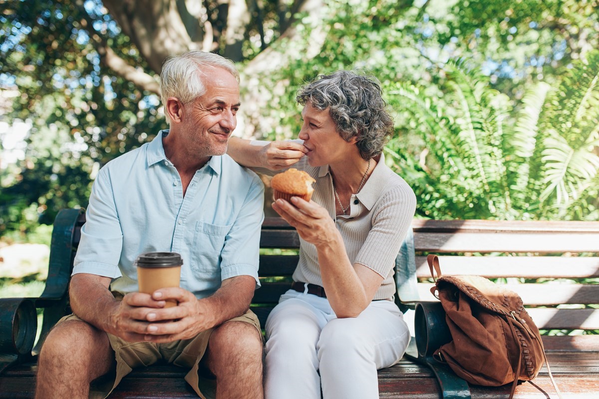 Older man and woman enjoying coffee and a muffin on park bench.