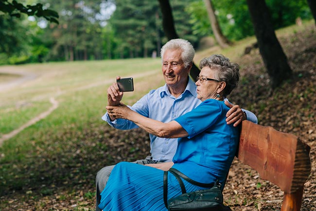 Older man and woman, sitting on park bench, taking a selfie.