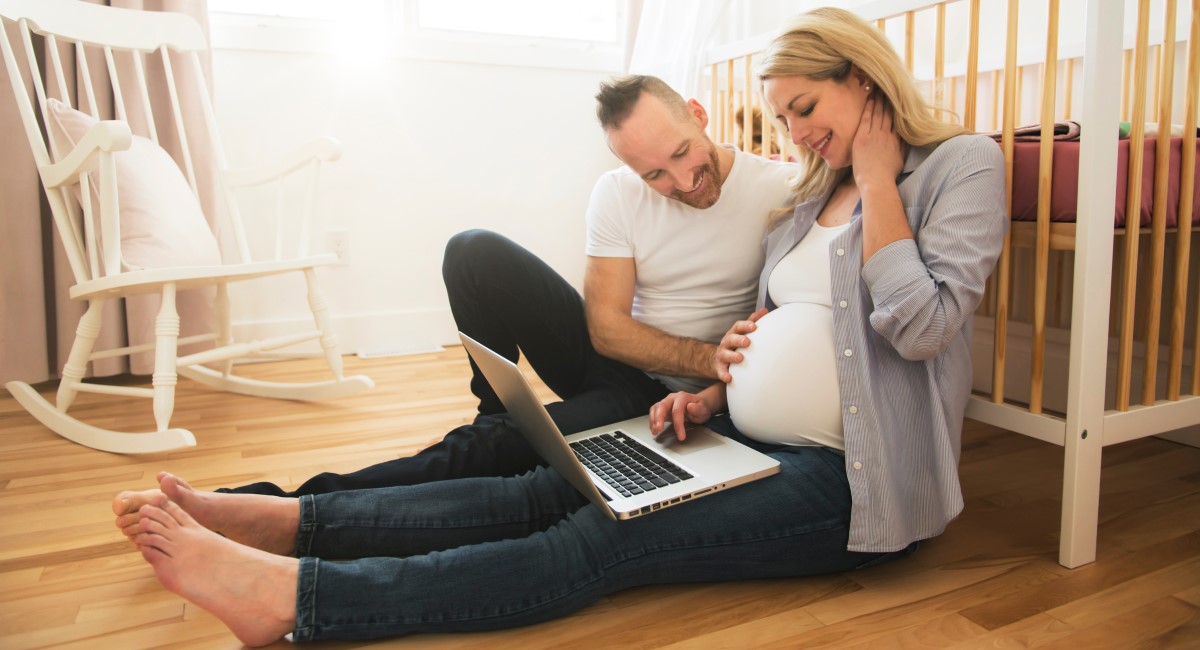 Pregnant woman and partner sitting on the floor, leaning against a new cot and looking at a laptop screen