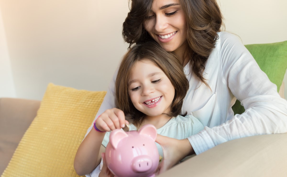 Smiling mother helping happy daughter put a coin in piggy bank