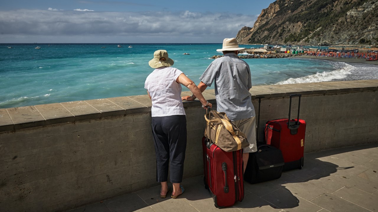 Older man and woman with suitcases looking out over a beach 