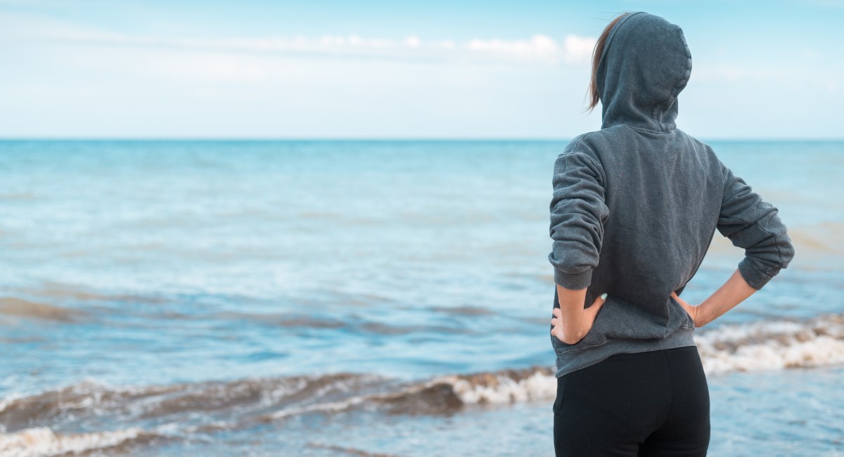 Woman in grey hoodie standing with back to camera, looking out over the ocean.