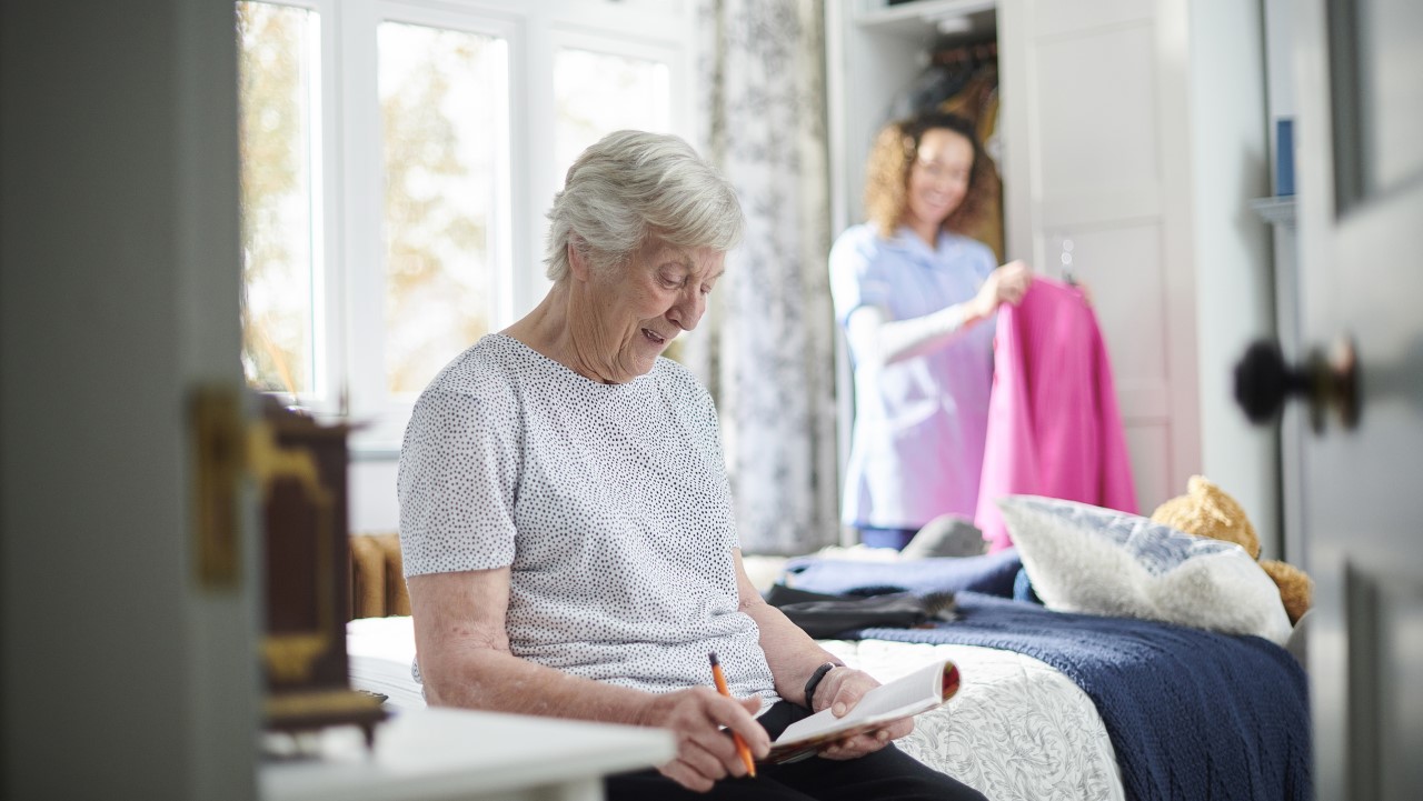 Older woman sitting on bed, with care worker in the background