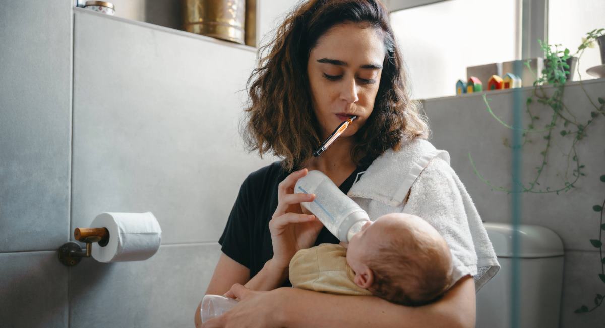 A woman brushing her teeth and feeding a baby at the same time