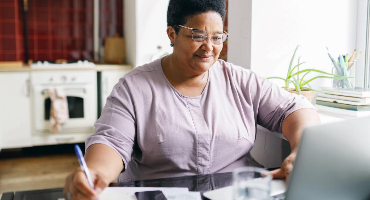 A woman sitting at a table in a kitchen smiling at a computer and writing a list