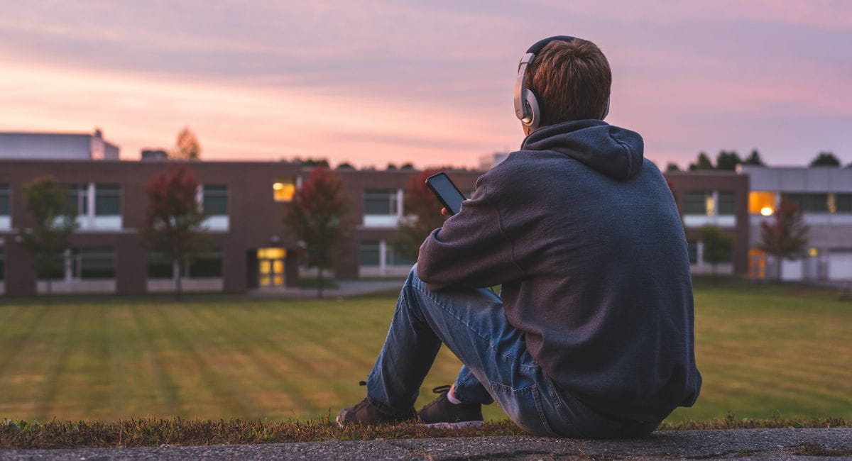A young man with headphones sitting on a field alone as the sun sets