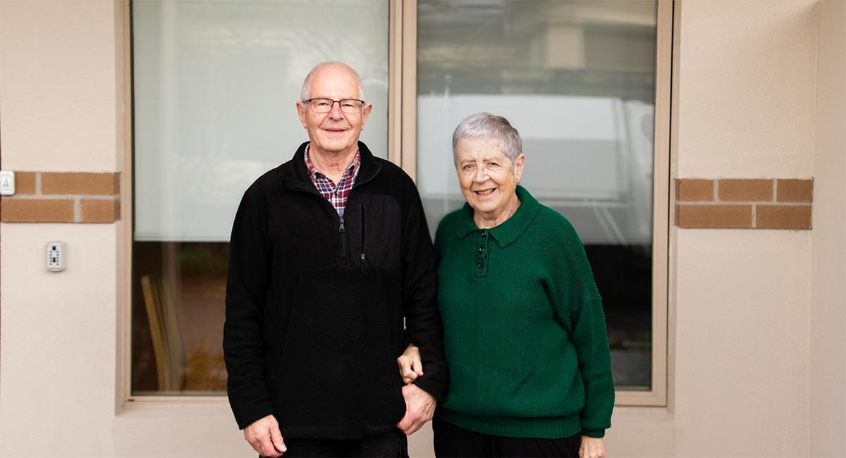 Older couple Barbara and Fred Seeber smiling at the camera in front of the window of their unit at Victoria Grange Retirement Community.
