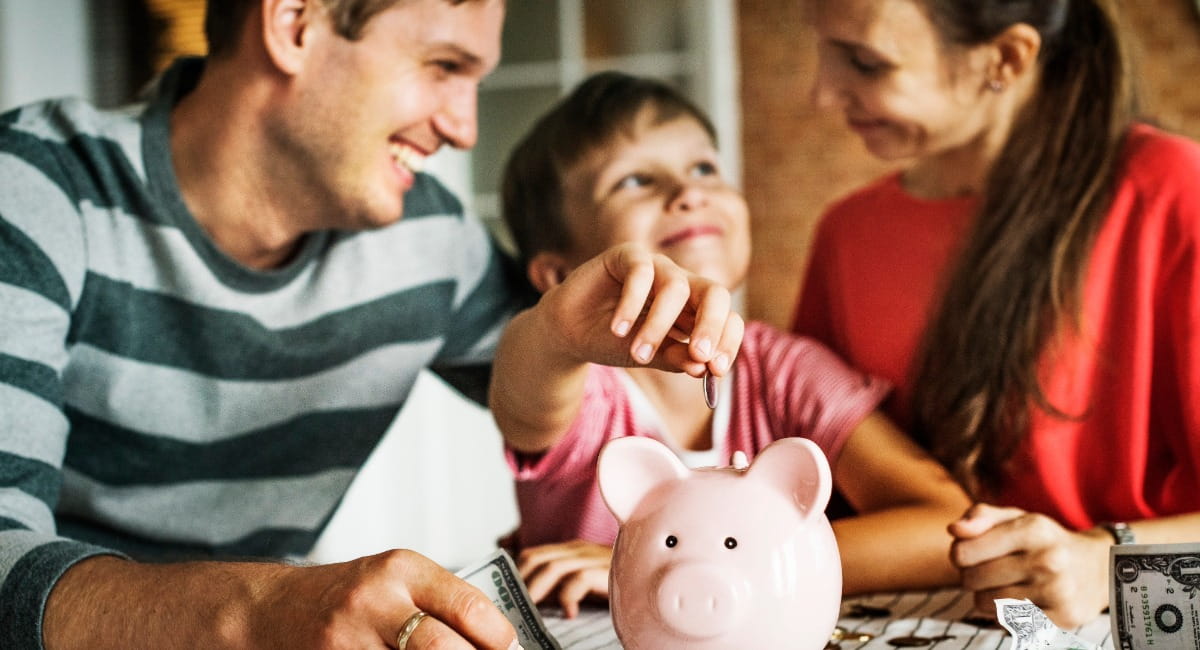 A young girl putting money in a piggy bank while her parents smile from either side of her