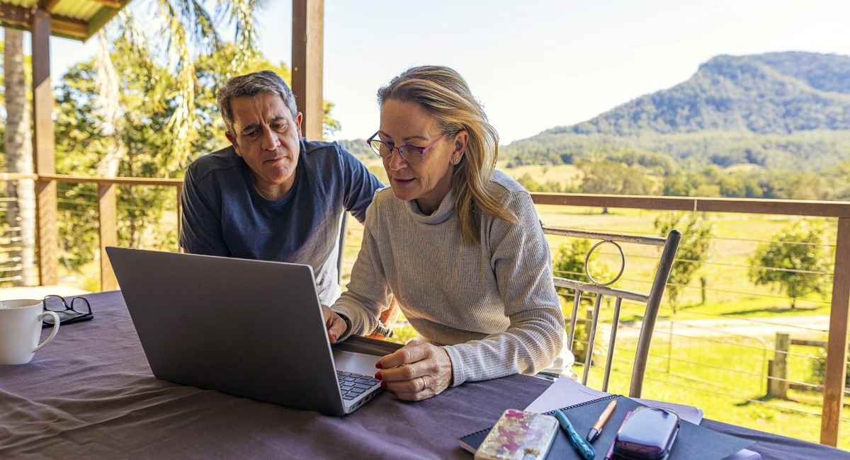 A middle aged woman and her partner sitting on a balcony with mountains in the distance while looking at a computer