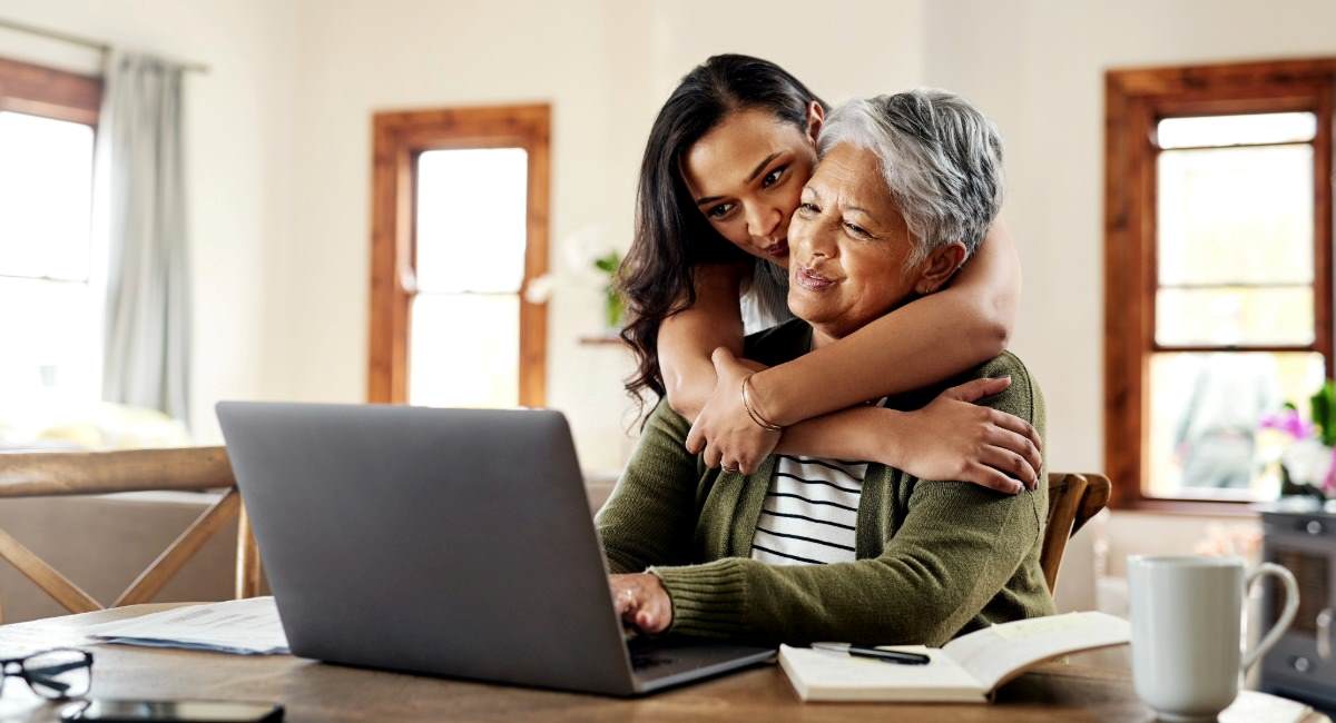 A senior woman sitting at a laptop while her daughter hugs her from behind