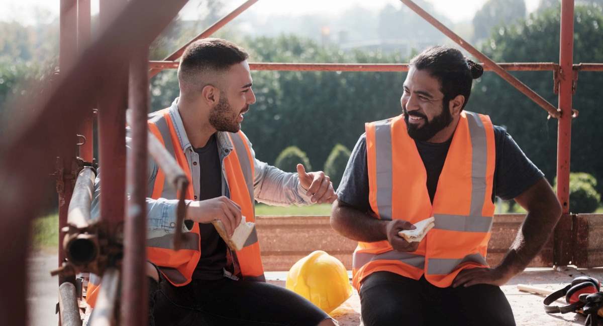 Two construction workers sitting onsite while eating sandwiches and laughing