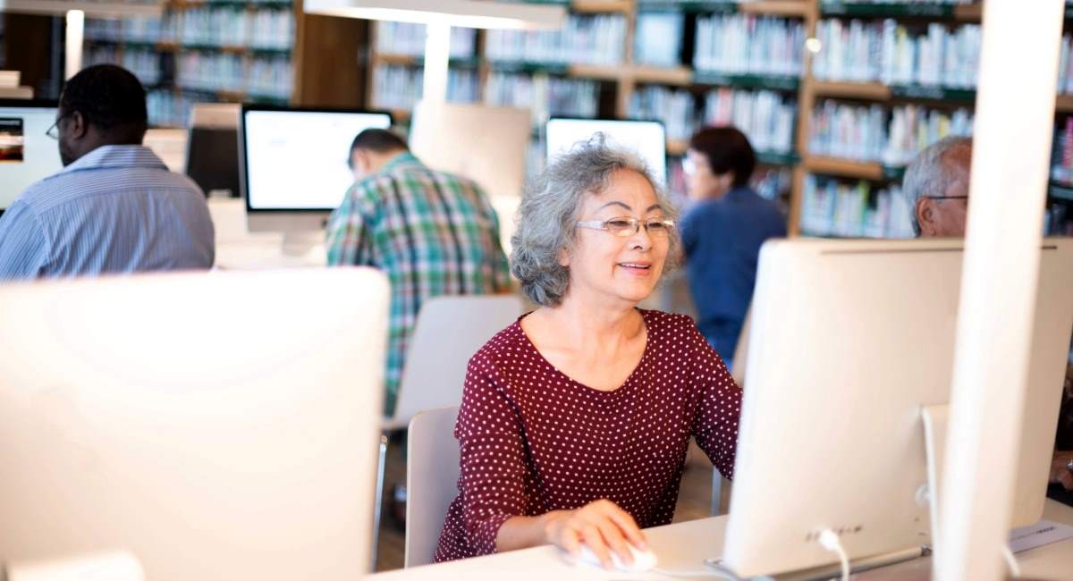 A mature woman smiling and sitting at a computer in a library surrounded by others on computers 