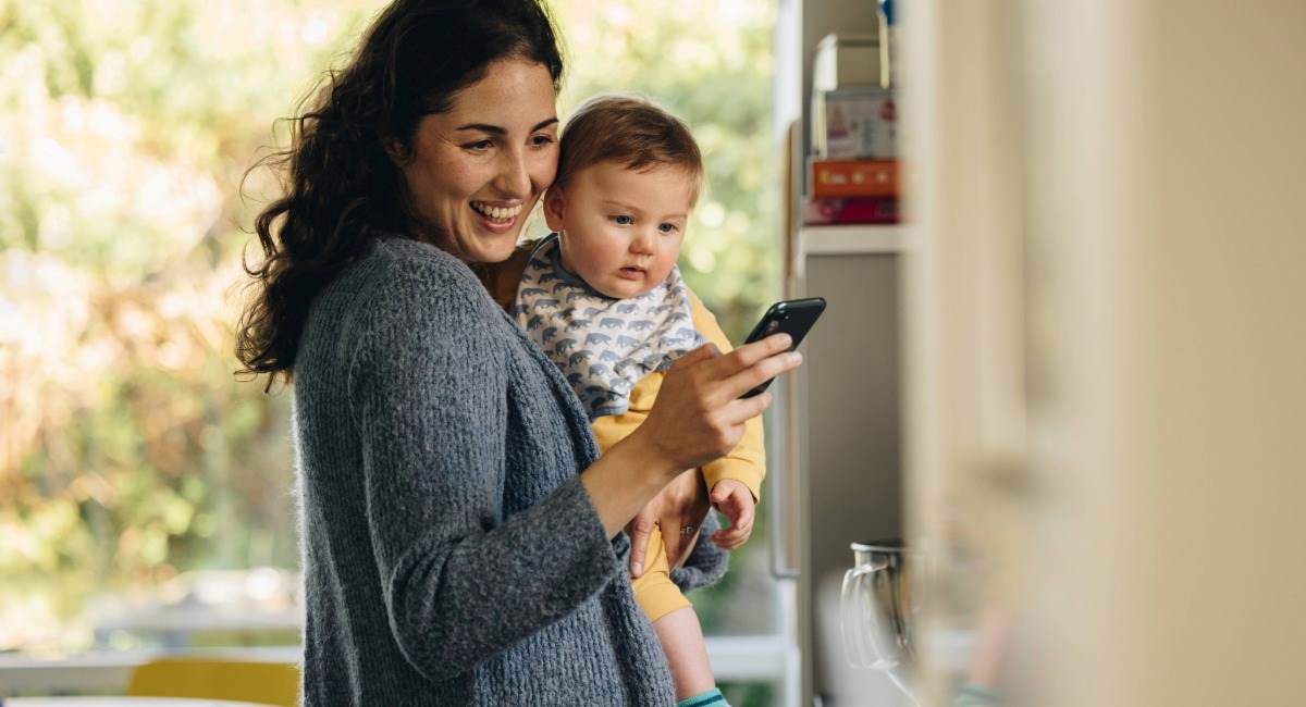A woman holding a baby while smiling widely at her phone