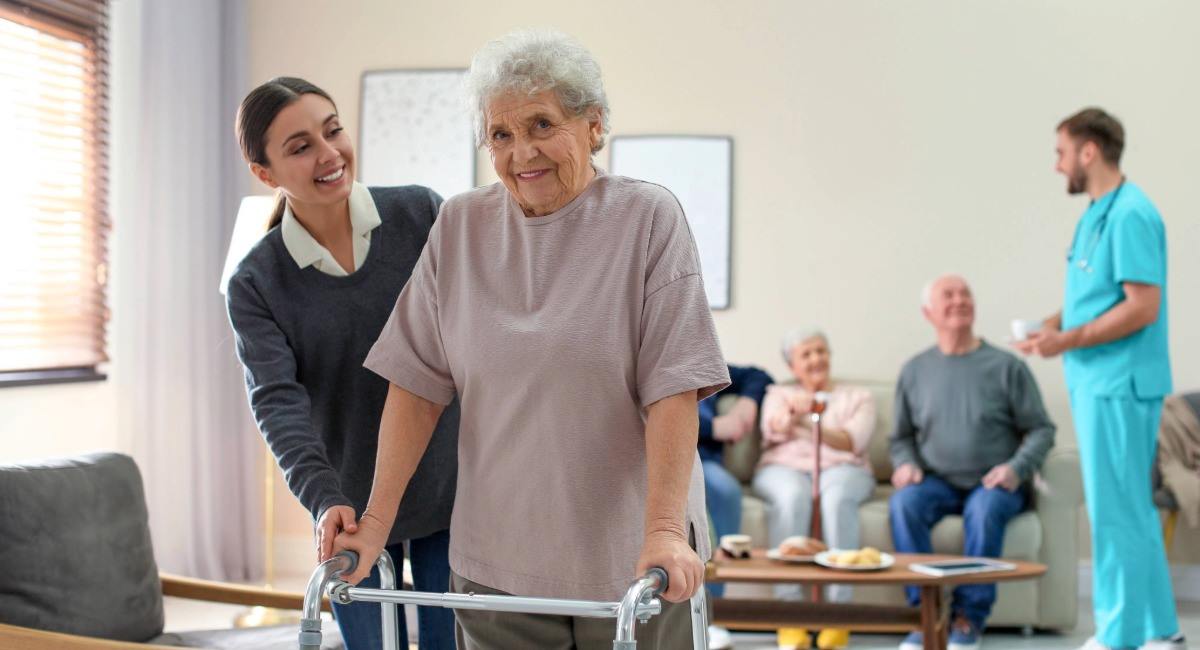 An elderly woman leaning on a walking frame and being assisted by a young caretaker