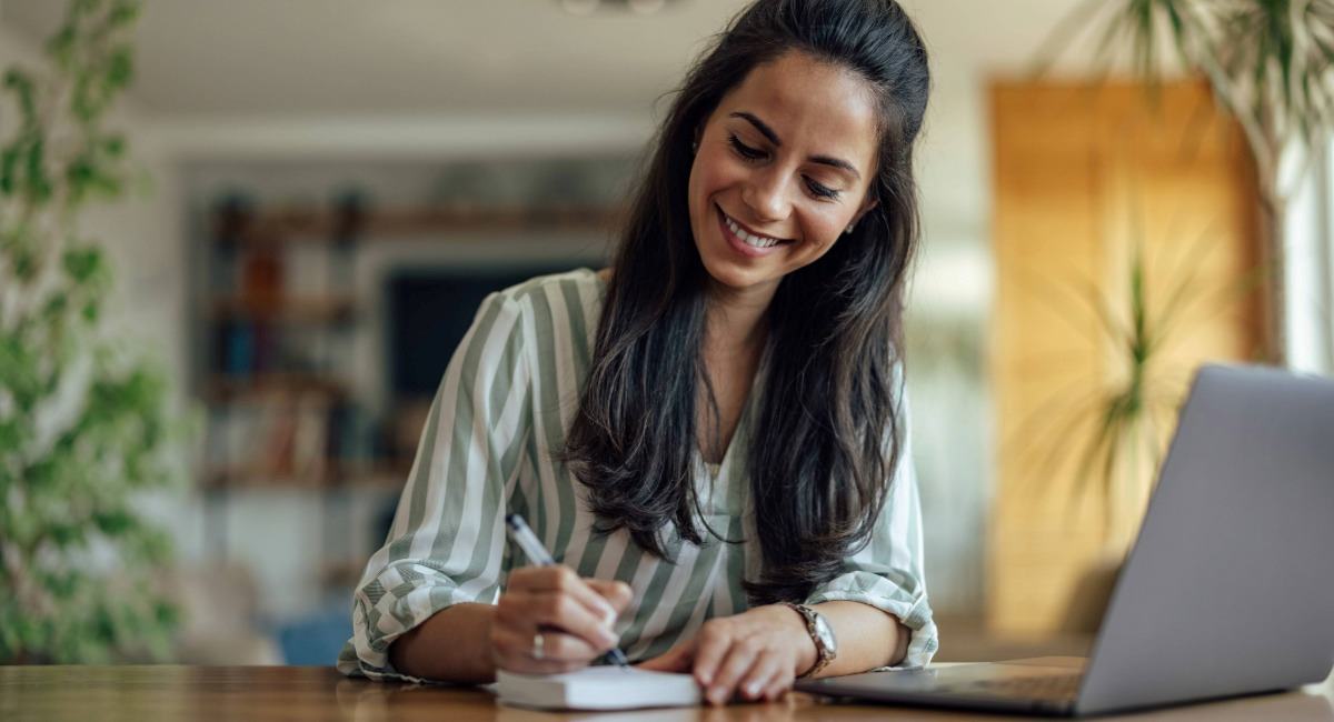 A young woman smiling while writing on a notepad in front of a computer