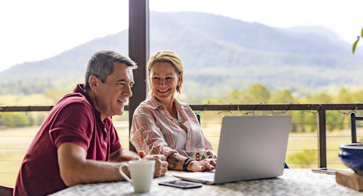 A couple in their 50s sitting on a balcony overlooking a mountain while smiling at a computer