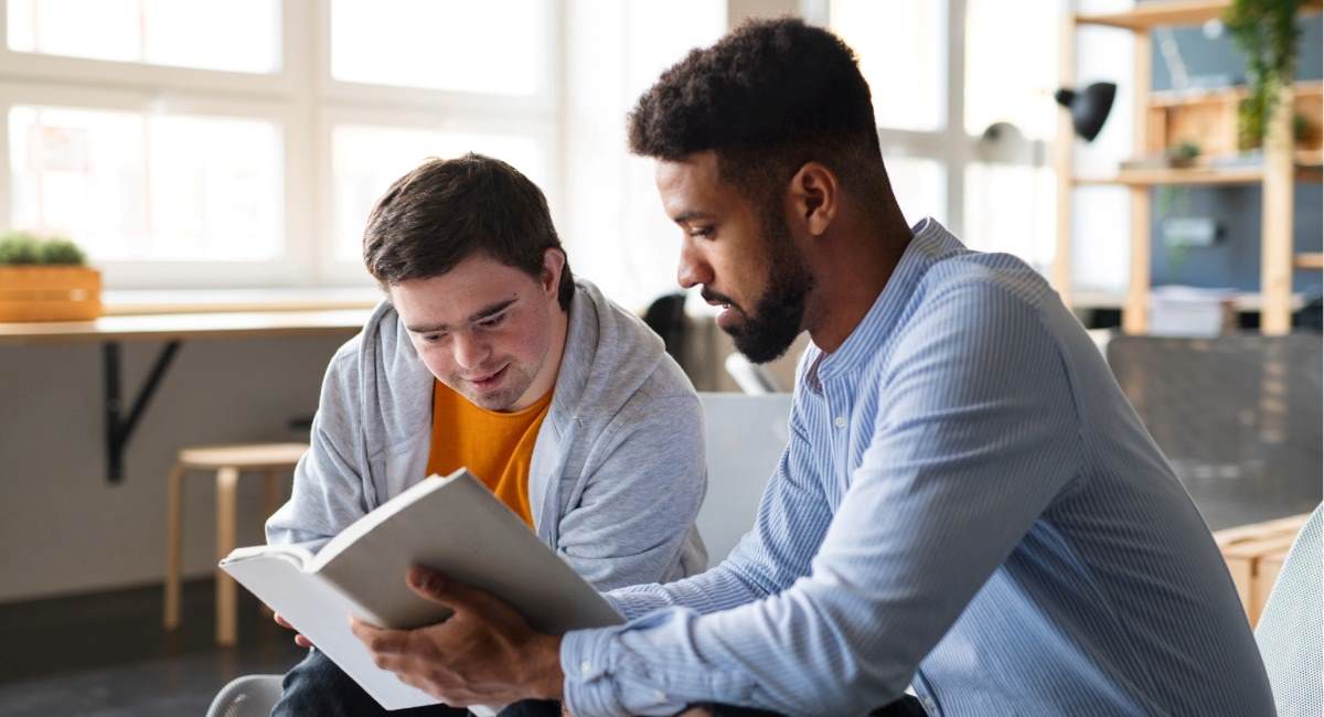Young happy man with Down syndrome and his tutor studying indoors at school