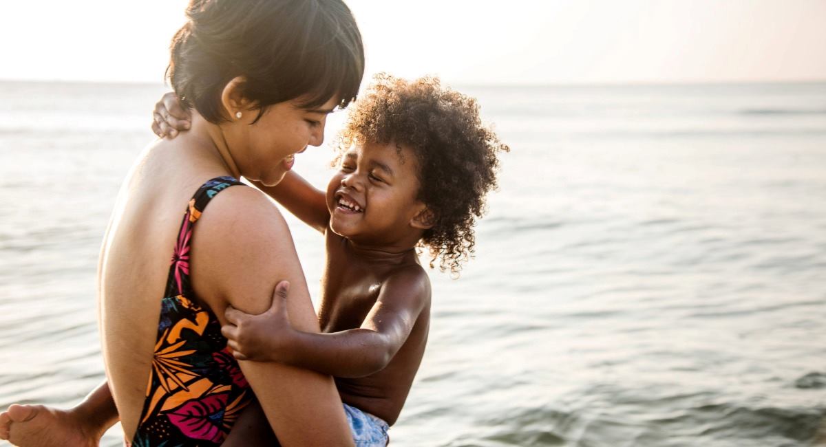 A woman holding a young child as they both smile at each other while standing in the ocean