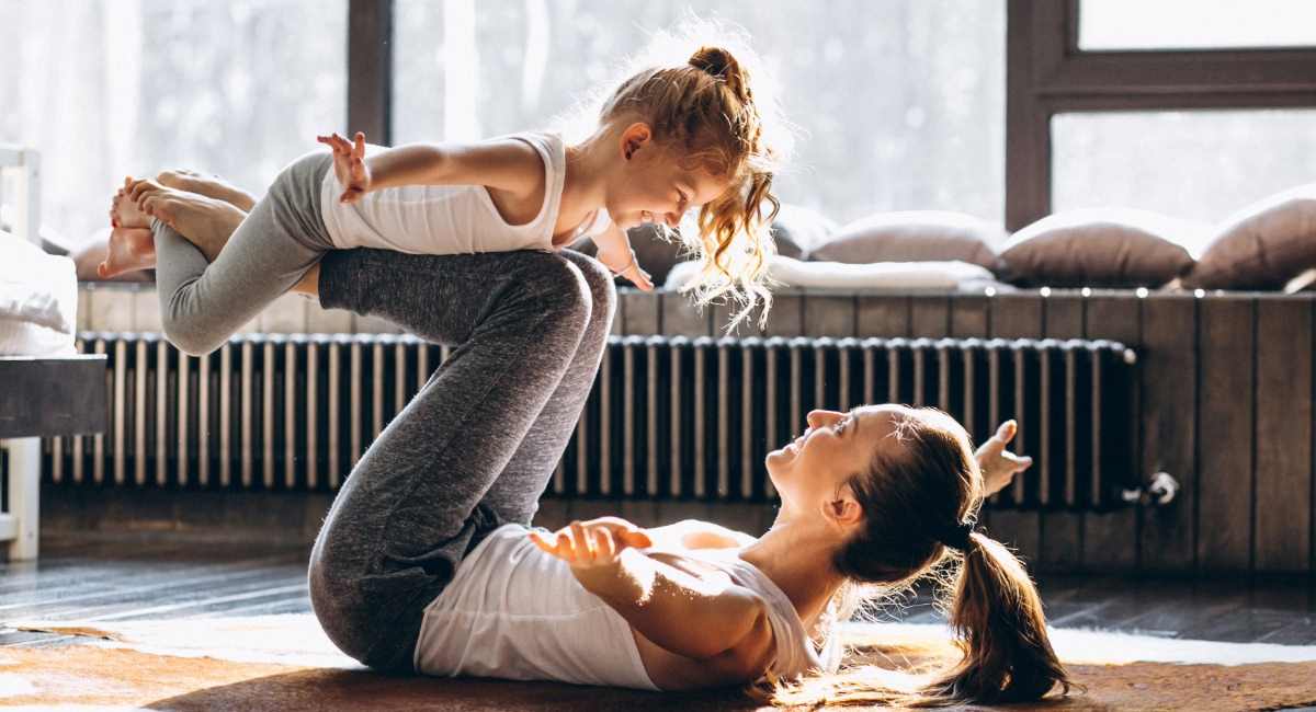 A woman in workout clothing on an exercise mat in a lounge room while playing with a young child