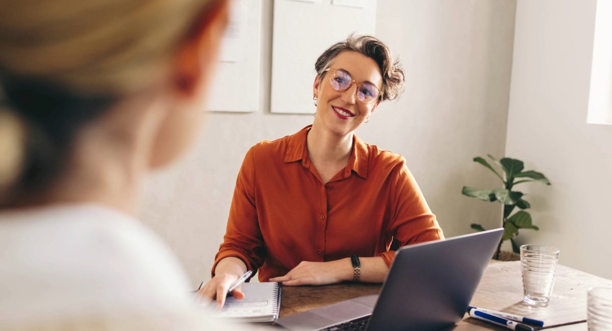 A manager sitting at a desk with a computer and notebook smiling at someone on the other side of the desk