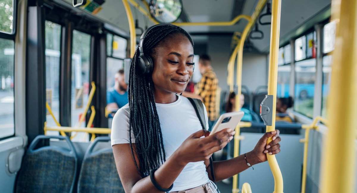 A young woman standing on the bus with headphones on, smiling at her phone