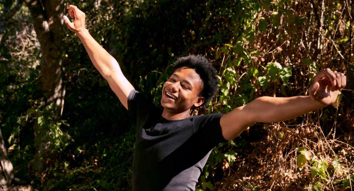 A young man smiling with his arms in the sky while standing near a tree