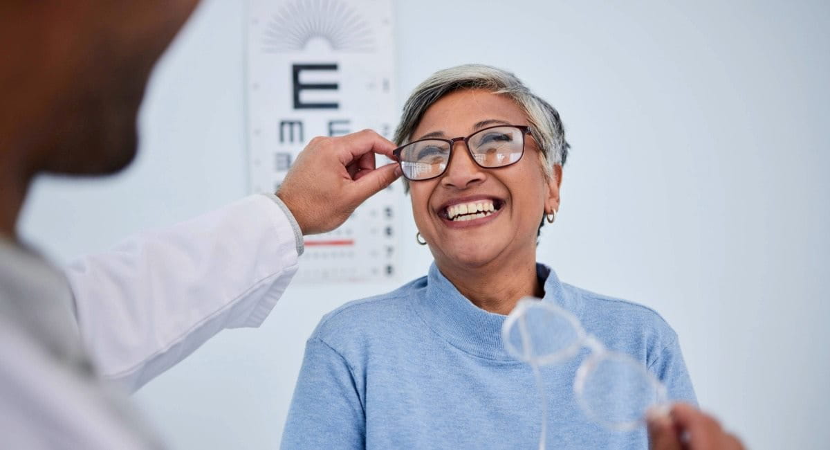 A woman in an optometrist office smiling while an optometrist puts glasses on her