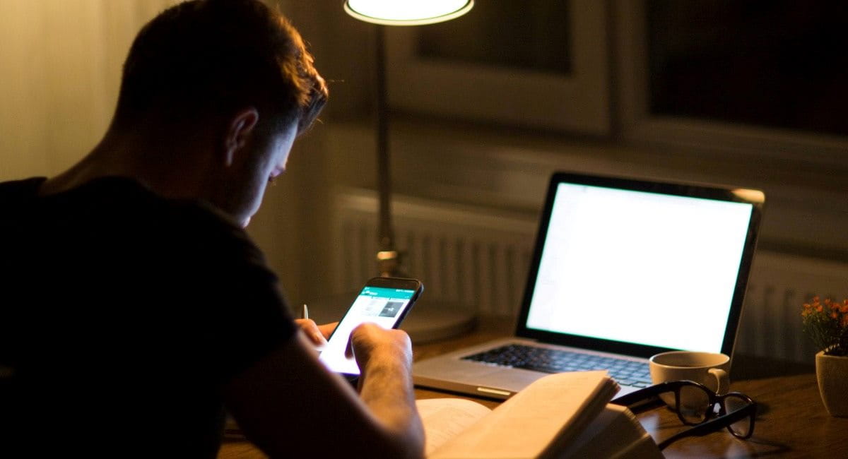 A man at a desk in the dark looking at his phone and laptop