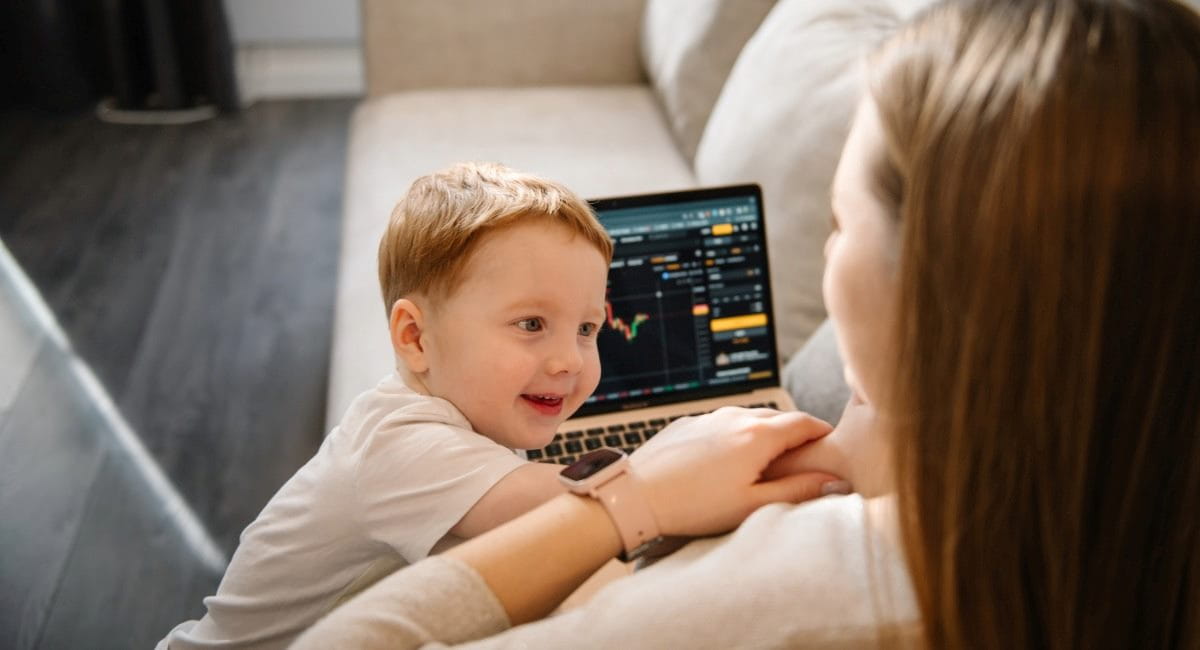 A woman sitting on a couch with a child while looking at investments on a laptop