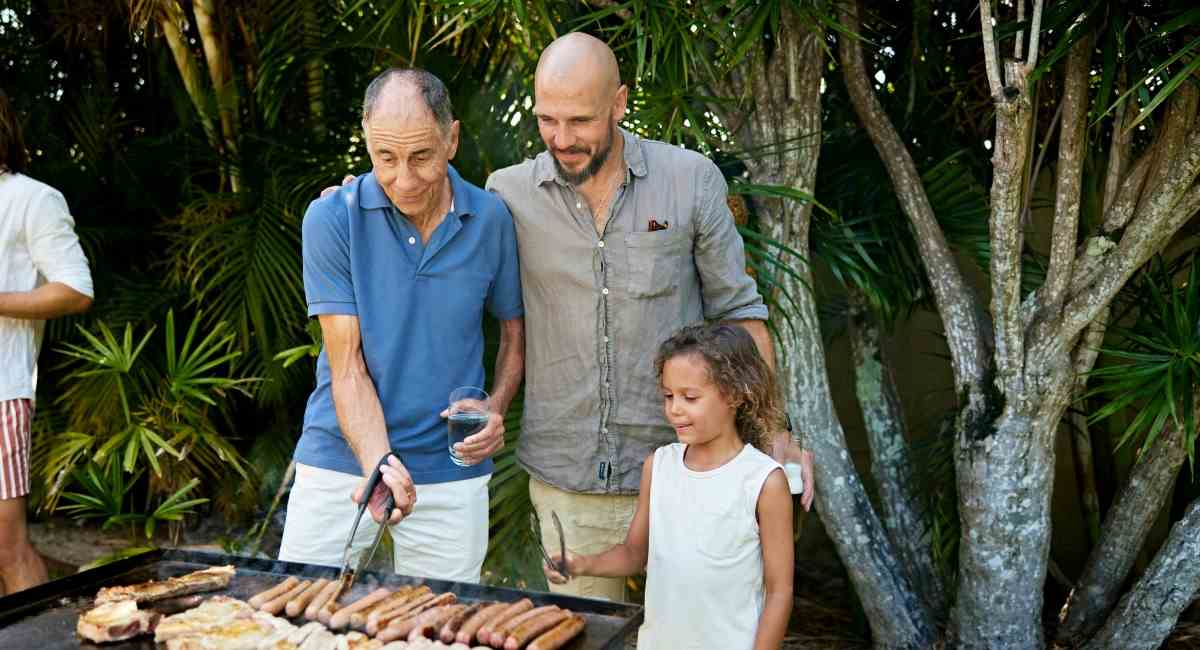 A middle aged man with his father and daughter cooking sausages on a barbeque