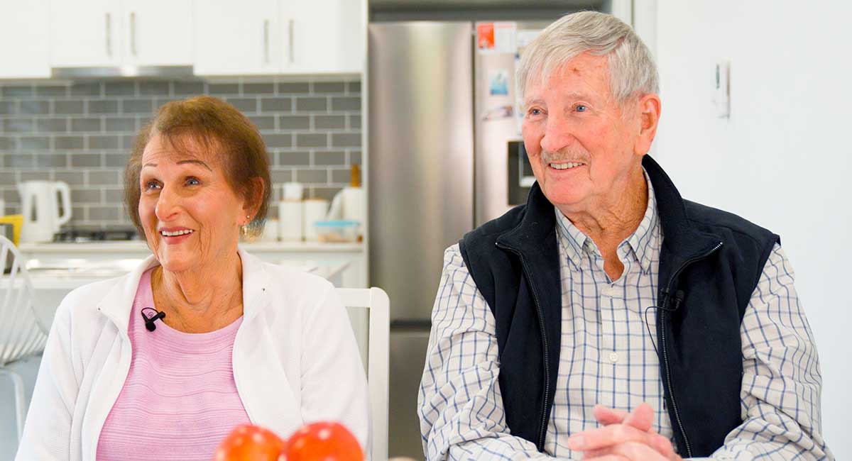 Image of older couple, Lynn and George, sitting together smiling in their kitchen.