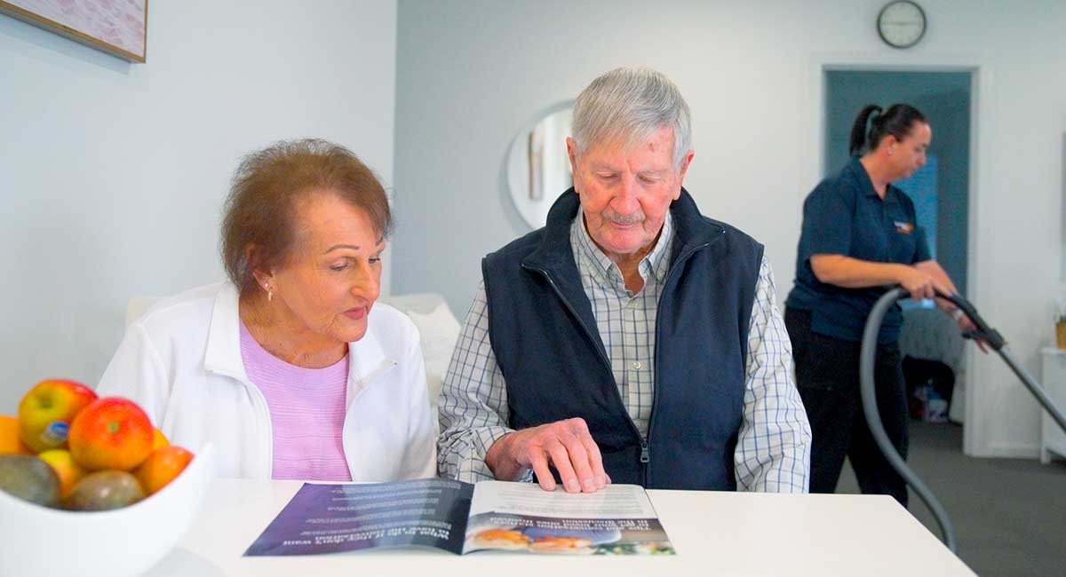 Older couple, Lynn and George, reading a magazine while a cleaner vacuums behind them.