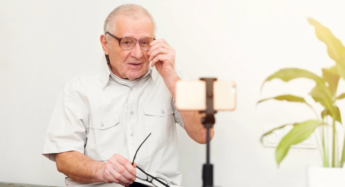 An elderly man wearing glasses sitting in front of a phone on a tripod
