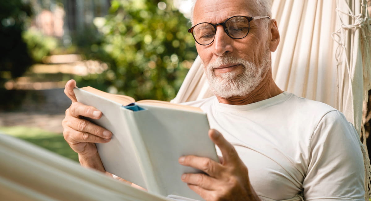 An older man reading a book on a hammock 