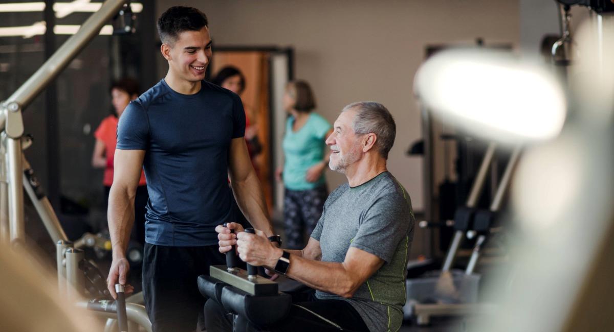 An older man using a machine at the gym while speaking to a young man
