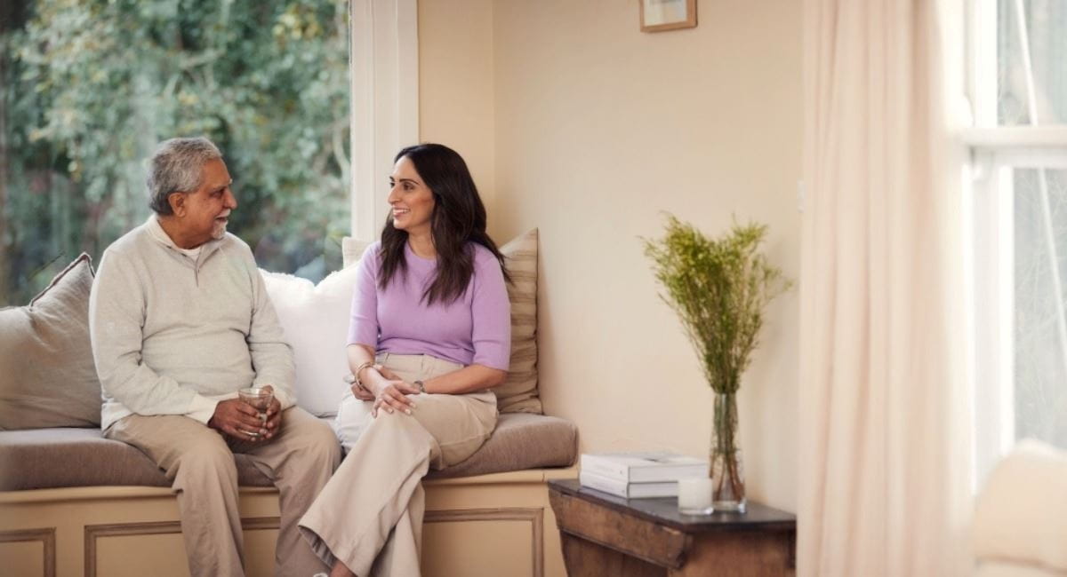 A middle aged woman and her father sitting by the window speaking to each other
