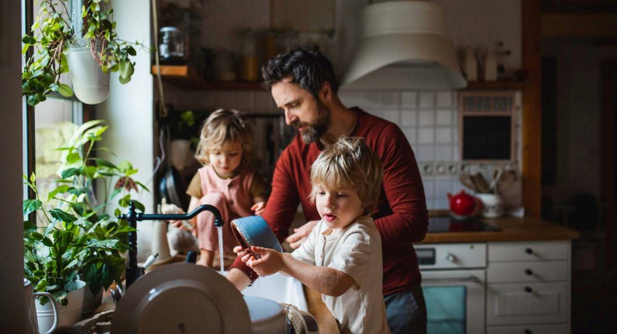 A man washing dishes in a kitchen with two young children