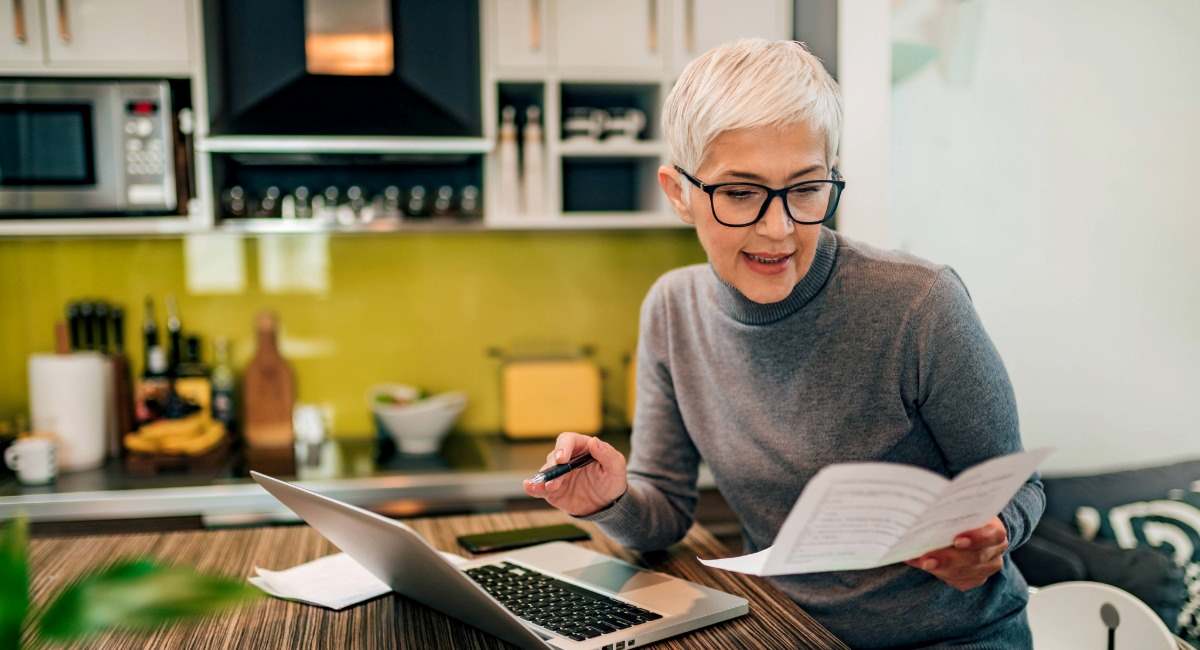 A woman sitting with a computer at a kitchen table looking at paperwork