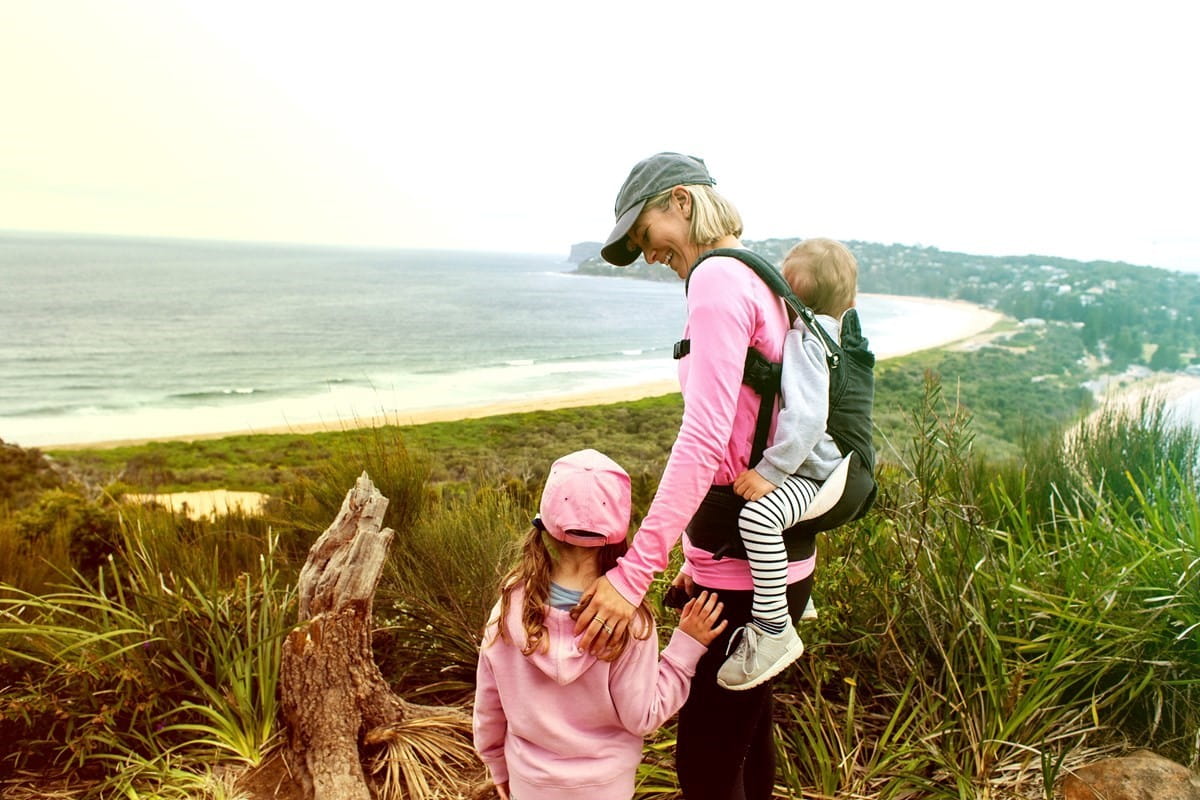 Happy mother with child at her side and baby in harness on her back, overlooking beach