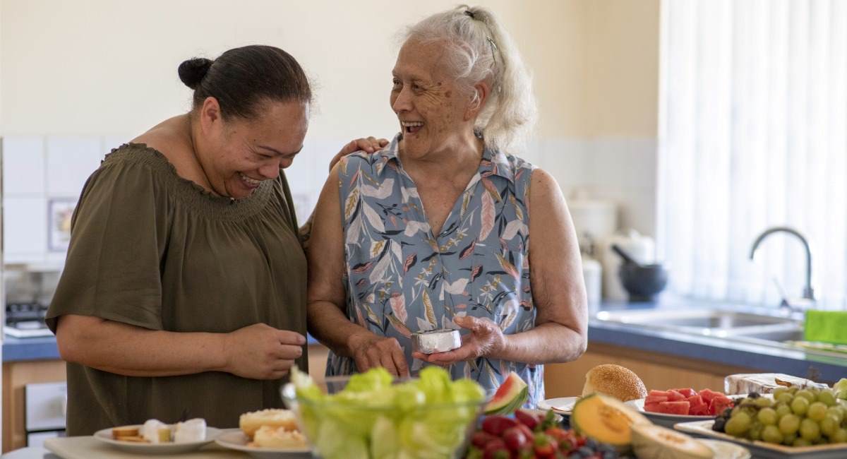 Two mature women laughing in a kitchen while preparing healthy food