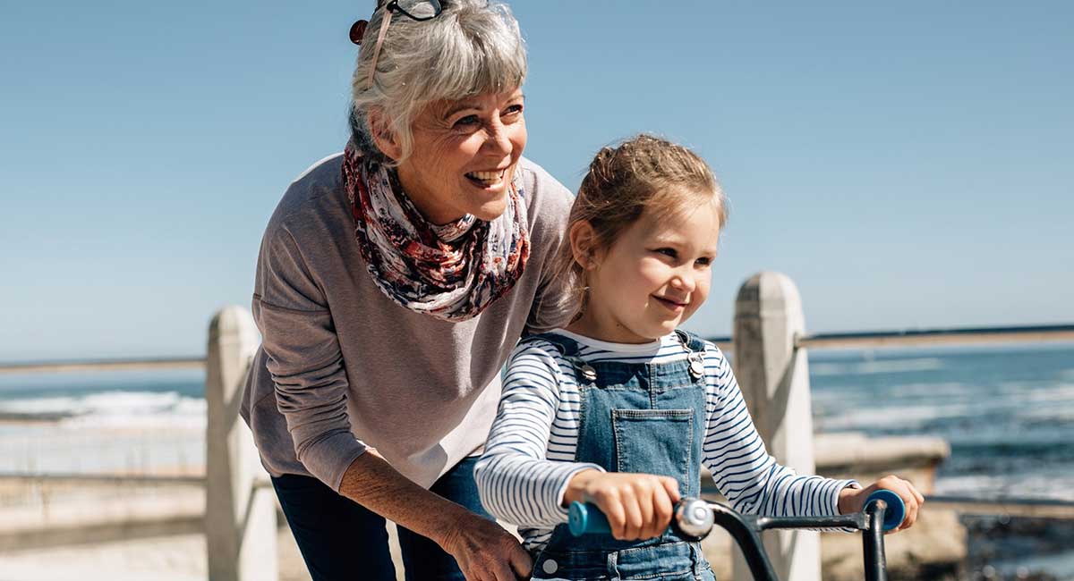 Grandmother with young granddaughter, pushing her while she rides a bike next to the beach on a sunny day.