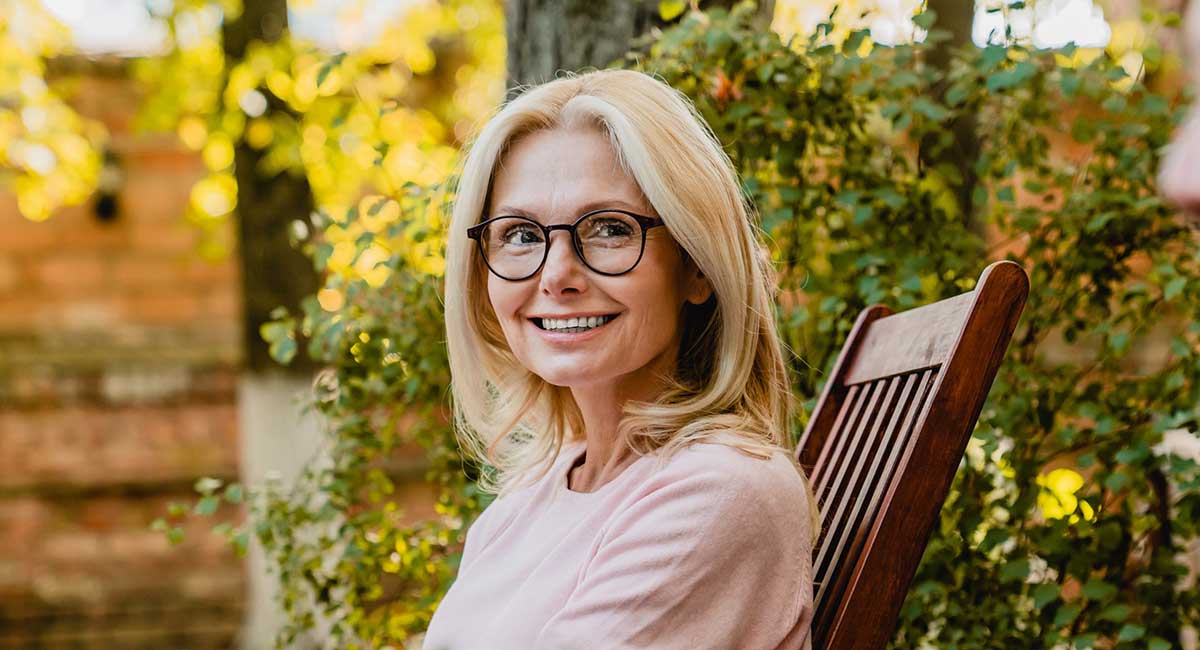 Blonde woman in early 50s with glasses, sitting on a garden chair surrounded by leaves starting to change in autumn.